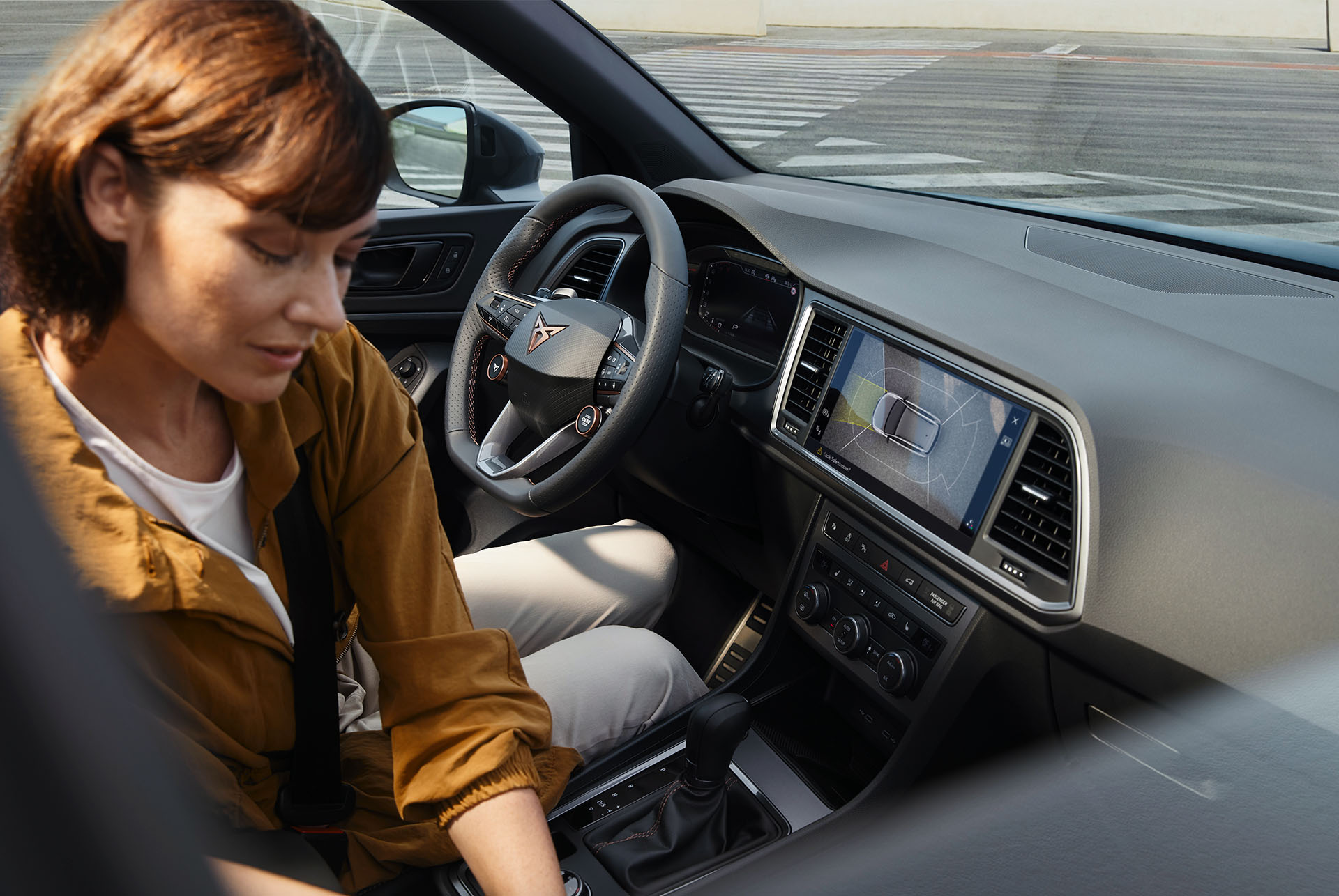 L'intérieur de la CUPRA Ateca met en évidence les caractéristiques technologiques avancées. Le tableau de bord affiche un grand écran d'infodivertissement avec navigation et le volant avec boutons satellites intègre des commandes. Une femme est assise à l'intérieur. 