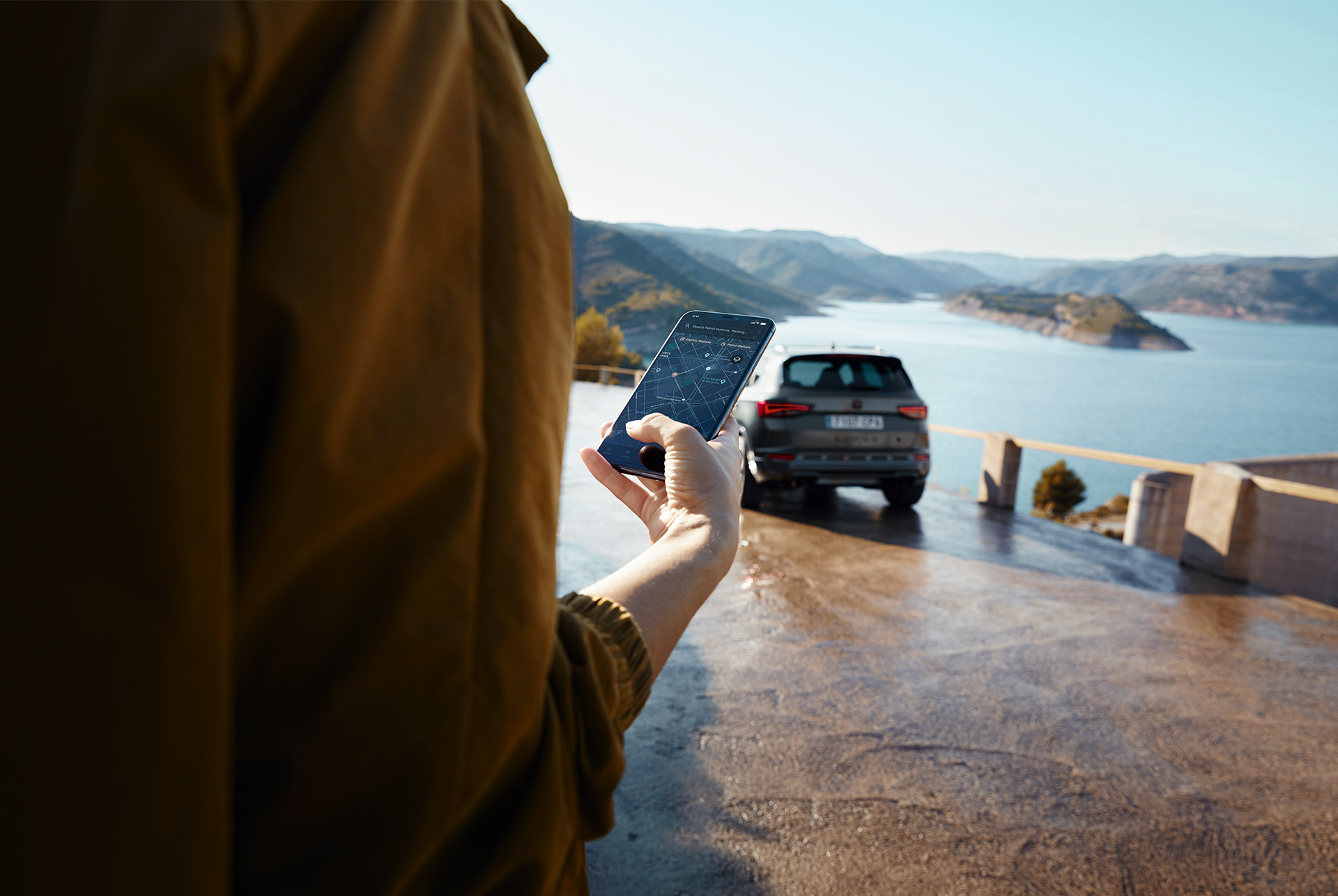 Une personne tenant un smartphone avec la CUPRA Ateca en arrière-plan, garée près d'une vue panoramique de montagnes et d'eau, symbolisant un monde connecté.