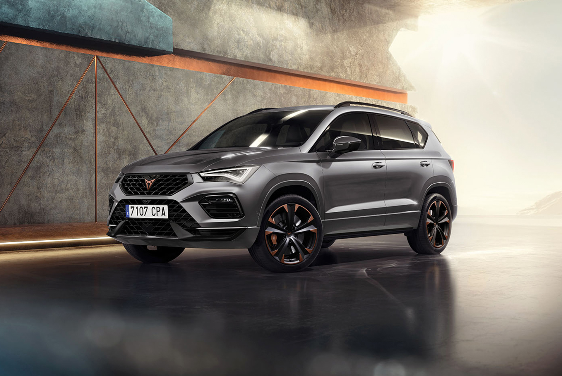 Une cupra ateca  en gris graphite, un SUV compact et sportif garé dans un cadre moderne et industriel. Le design de la voiture se caractérise par des lignes aérodynamiques, des phares à LED et des jantes en alliage distinctives aux accents cuivrés. L'arrière-plan comprend un mur géométrique qui contraste avec le design épuré du véhicule.